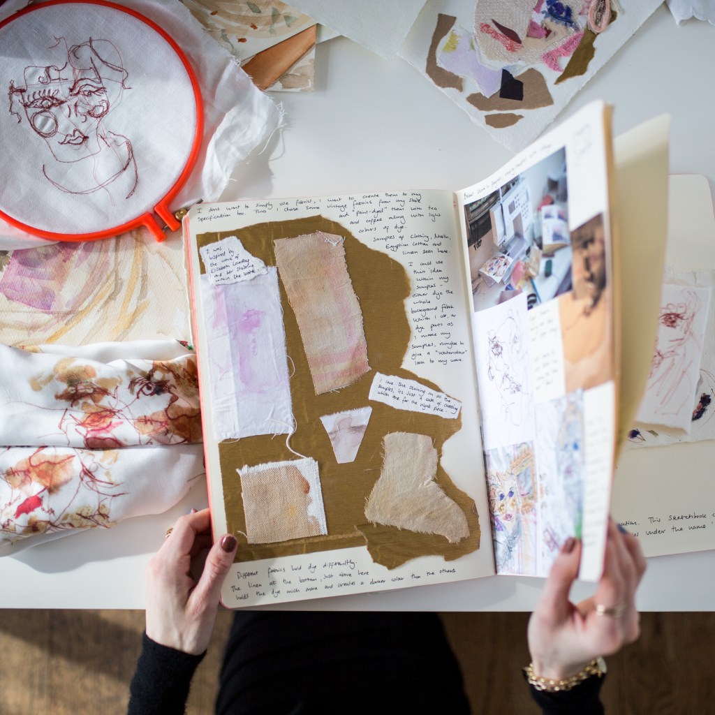 hands turning a sketchbook page, surrounded by fabric on a table